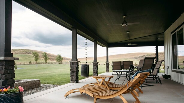 Un patio couvert offrant une vue sur un terrain de golf, avec des fauteuils de jardin en bois, une table et des piliers en pierre.