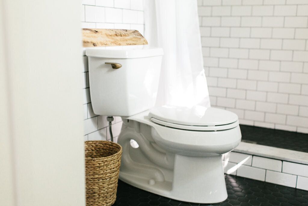 A white toilet with a wooden shelf above it, a wicker basket on the floor, and a black tiled floor.
