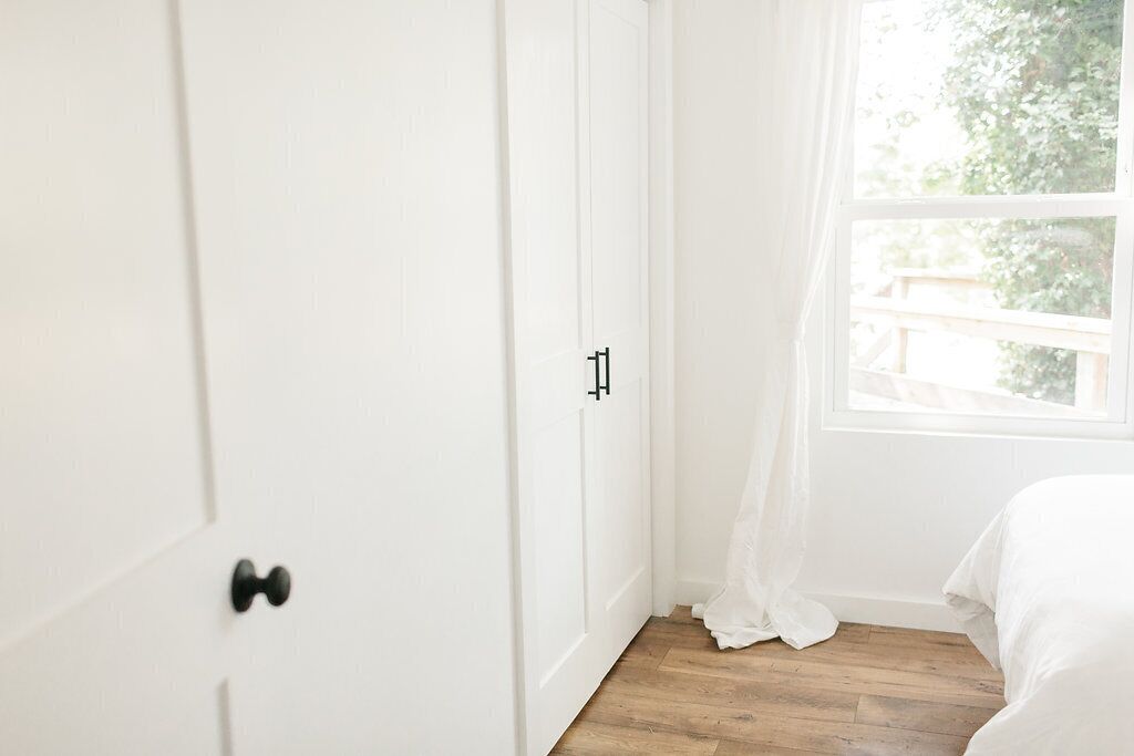 Une chambre avec un plancher en bois, des murs blancs, un lit avec une literie blanche et une fenêtre ornée de rideaux légers.