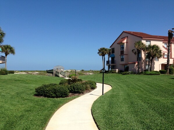 A well-maintained grassy area with a curved pathway leading to a building with multiple balconies and a red-tiled roof. - Ocean front Condo ground level REMODELED (St. Augustine)