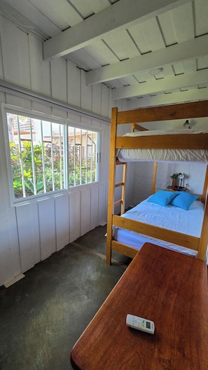A bunk bed with a wooden frame and blue pillows, a bedside table with a remote control, and a window with bars and greenery outside.