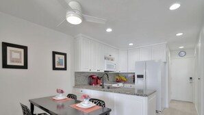 A modern kitchen with white cabinets, a microwave, a refrigerator, and a dining area with a table and chairs. - Newly Remodeled! Oceanfront Mission Beach Condo Sleeps 6 with Incredible Views! (San Diego)
