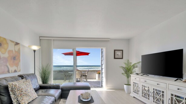 A living room with a leather sofa, a coffee table, a flat-screen TV, and a balcony with a red umbrella. - Newly Remodeled! Oceanfront Mission Beach Condo Sleeps 6 with Incredible Views! (San Diego)