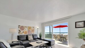 A modern living room with a leather sofa, a coffee table, and a large window offering a view of the beach. - Newly Remodeled! Oceanfront Mission Beach Condo Sleeps 6 with Incredible Views! (San Diego)