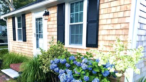 Een huis met een veranda, een raam met zwarte luiken en een tuin met blauwe hortensia's.