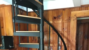Een wenteltrap met houten treden en zwarte metalen leuningen, in een ruimte met houten wanden en een open haard van baksteen.