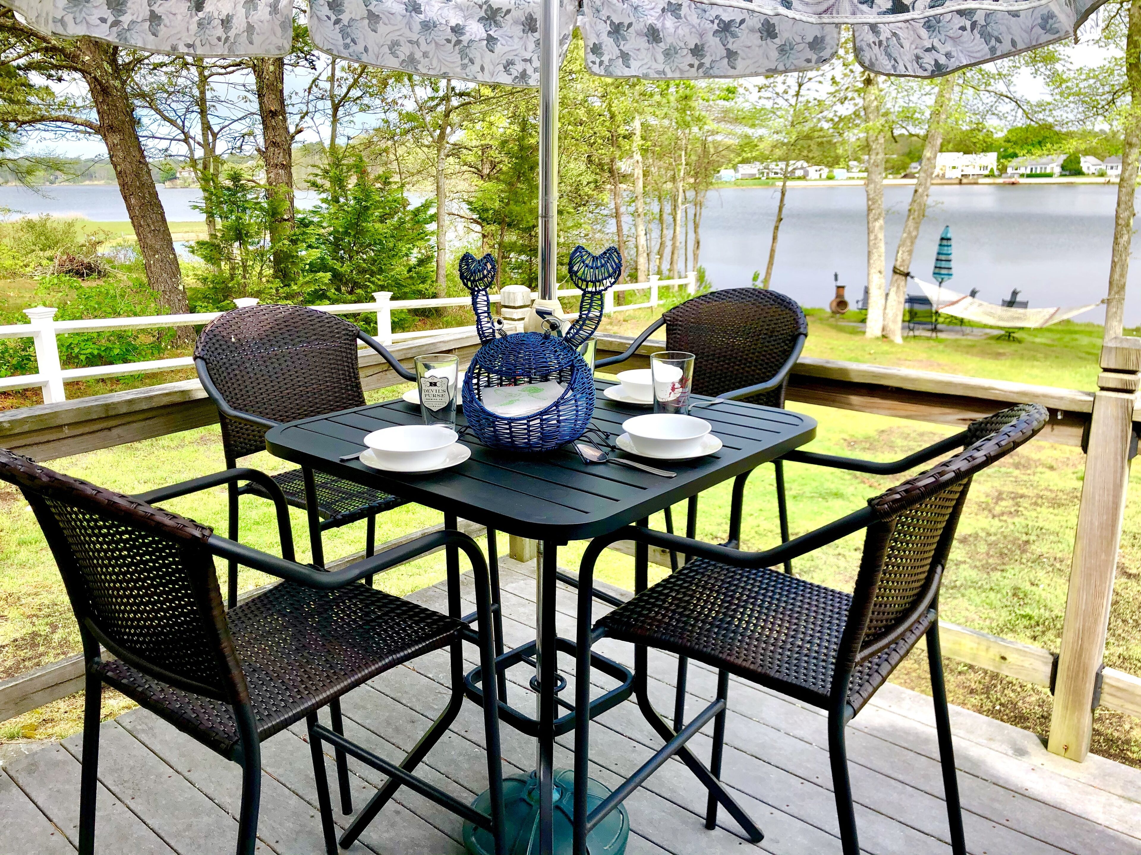 Uma área de refeições ao ar livre com uma mesa posta para quatro pessoas, rodeada por cadeiras de vime, uma tigela decorativa azul e um castiçal.