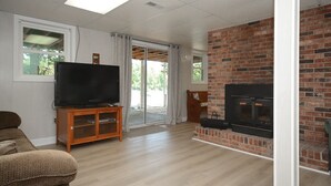 Een woonkamer met een televisie op een houten standaard, een open haard van baksteen en een schuifdeur naar een terras.