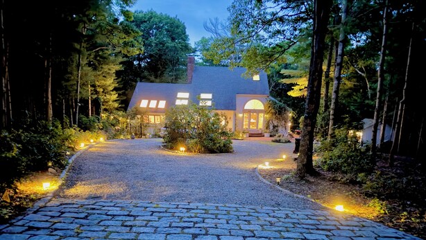 A house with a gravel driveway leading to a front door, surrounded by trees and illuminated by ground lights.