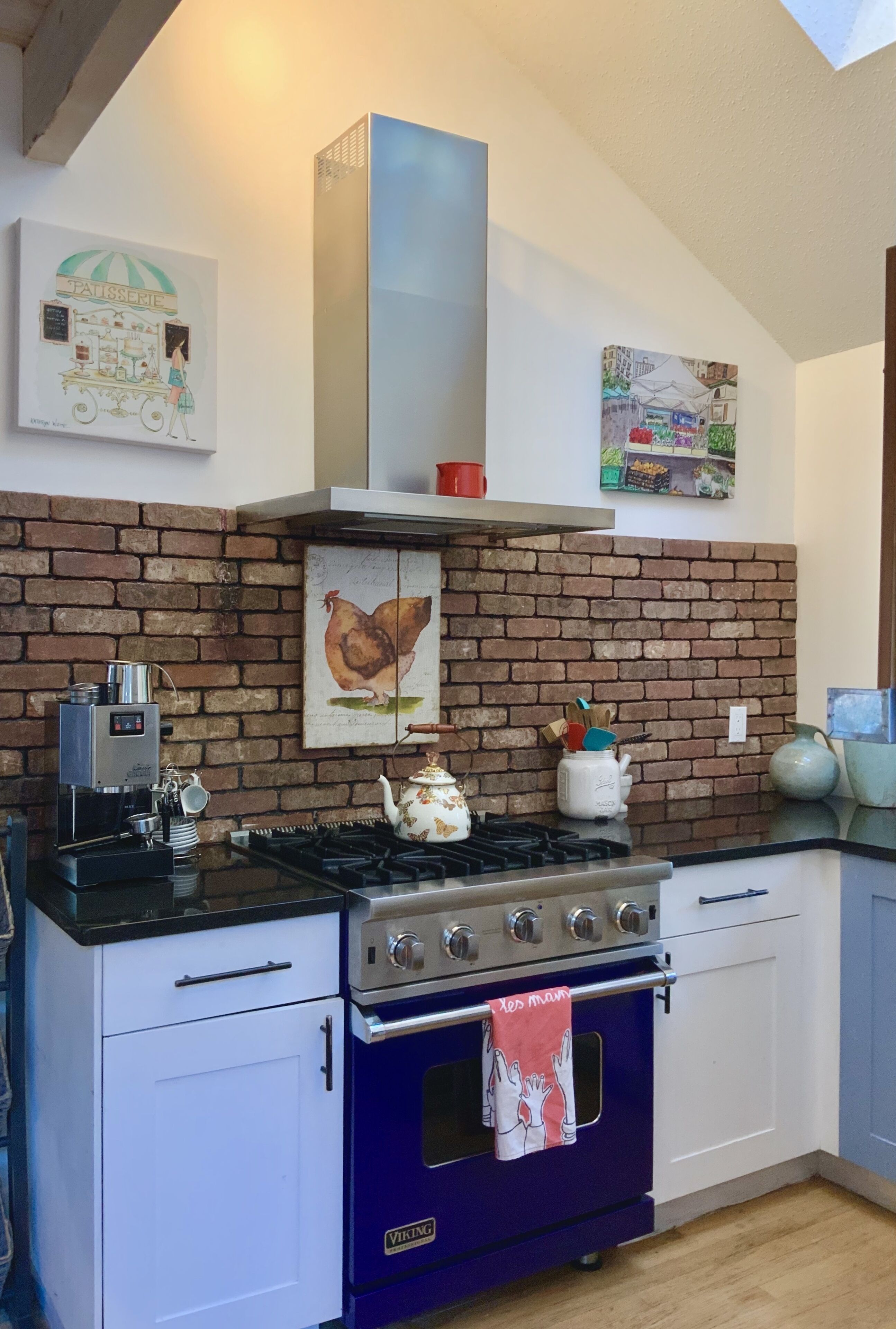 A kitchen with a brick wall, a gas stove, a blue oven, and white cabinets.