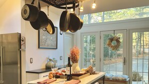 A kitchen with hanging pots, a refrigerator, a dining table with wine glasses, and a view of a garden through large windows.