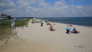 A beach with people sunbathing and swimming, a grassy area with a sign, and a clear sky.