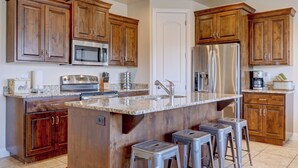 Una cocina con gabinetes de madera, electrodomésticos de acero inoxidable, encimera de granito y taburetes de bar.