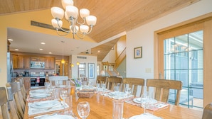 Un espace repas avec une longue table en bois dressée pour le repas, un lustre et un escalier menant à un niveau supérieur.