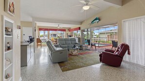 Amplia sala de estar con un sofá gris, un sillón rojo y una mesa de centro. Hay un ventilador de techo, una zona de cocina al fondo y vistas al exterior.