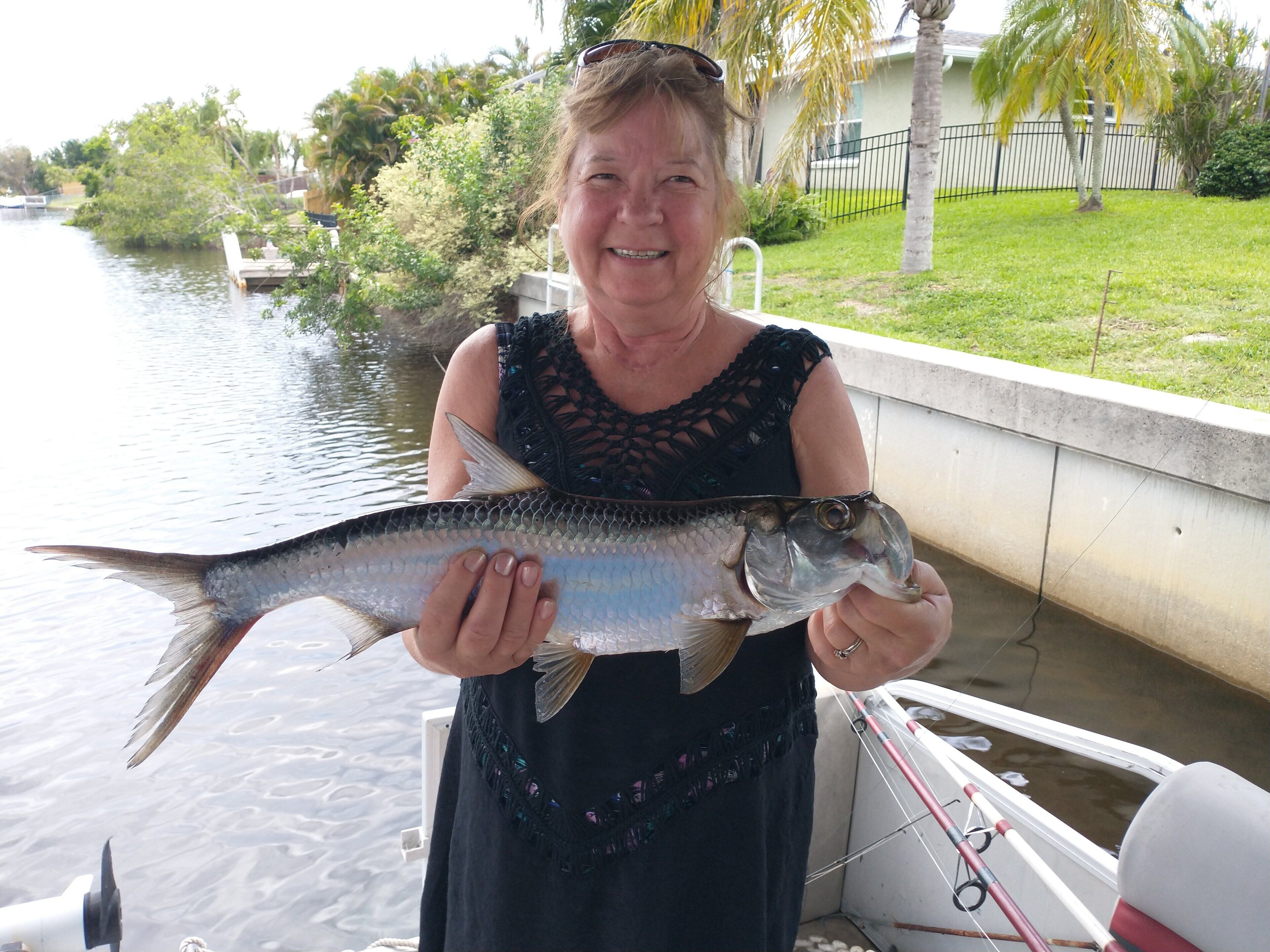 A person holding a fish near a water body with a boat and a dock in the background.
