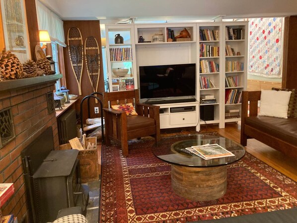 A cozy living room with a fireplace, wooden furniture, a TV, and a bookshelf filled with books. - Cottage at Taughannock: State Park, Cayuga Lake, Ithaca, & Cornell (Trumansburg)
