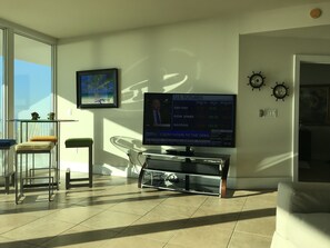Een moderne woonkamer met een grote flatscreen televisie waarop financieel nieuws wordt getoond, een glazen tafel met stoelen en uitzicht op de omgeving.