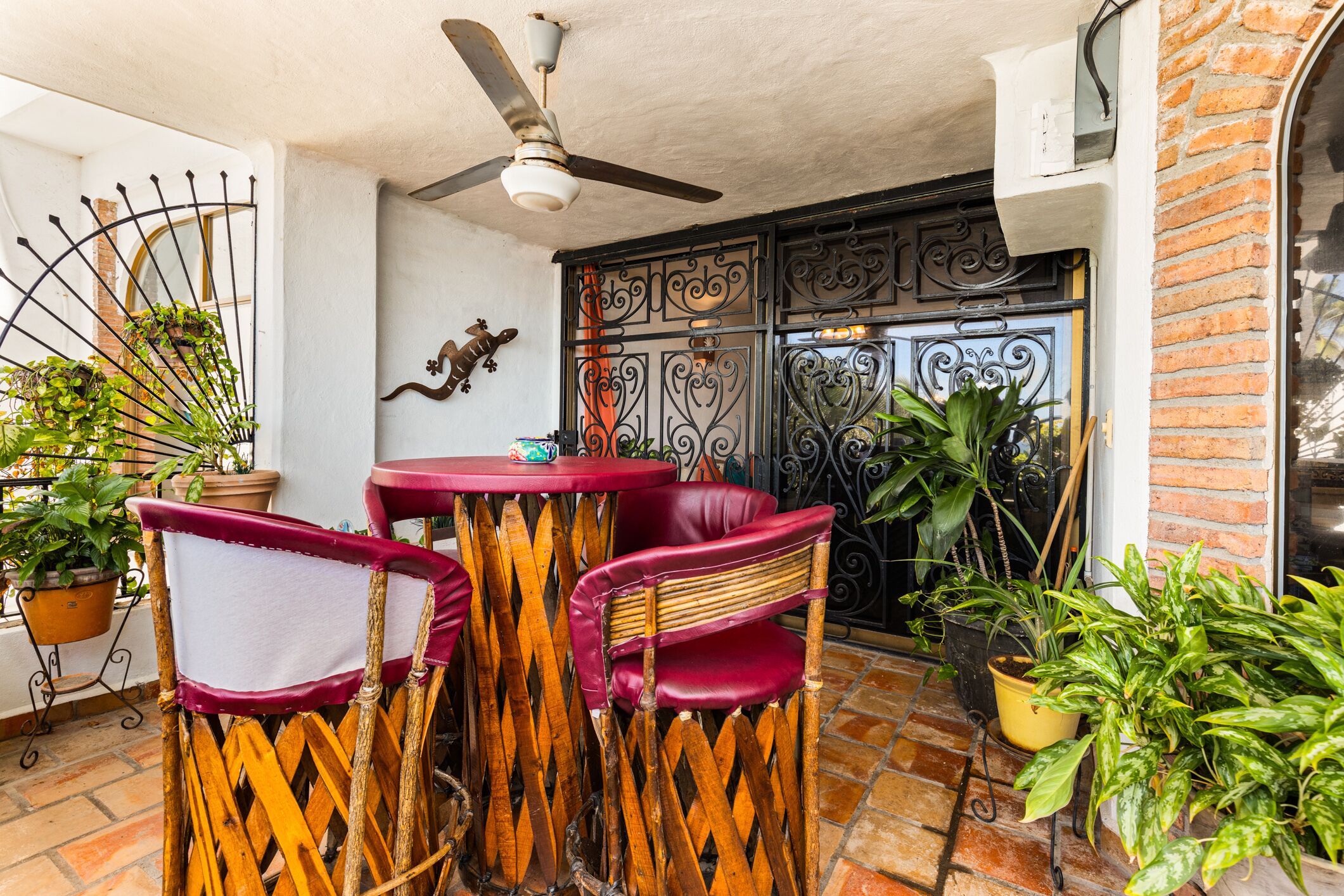 An outdoor seating area with wicker chairs and a red table, surrounded by potted plants and a decorative metal gate.