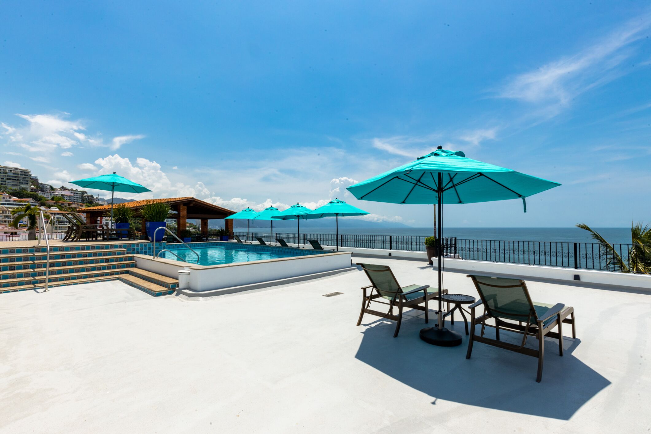A rooftop pool area with multiple turquoise umbrellas, lounge chairs, and a view of the ocean.