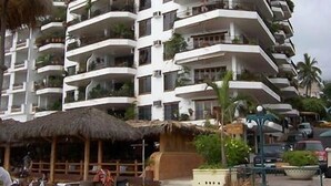 Um hotel com vários andares, varandas, uma construção com telhado de palha e um espaço pavimentado com assentos.