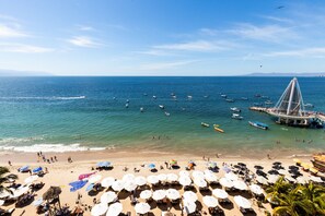 Una playa con numerosos sombrillas y tumbonas, un muelle con barcos y una vista despejada del mar.