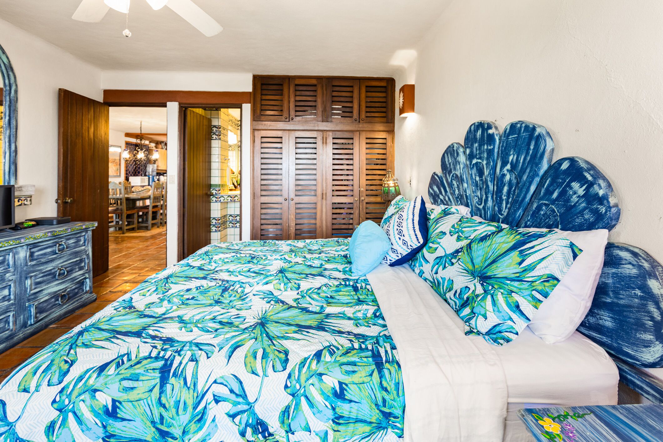 A bedroom with a bed featuring a tropical leaf pattern bedspread, a wooden headboard, and a white comforter.