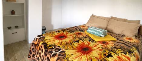 A bedroom with a bed covered in a floral-patterned bedspread, a wooden nightstand with a lamp, and a white wall with a shelf holding a framed document and a small box.