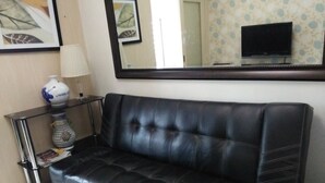 Ein schwarzes Ledersofa in einem Raum mit Fliesenboden, einem kleinen Tisch mit Lampe und Blumenvase sowie einem an der Wand montierten Flachbildfernseher.