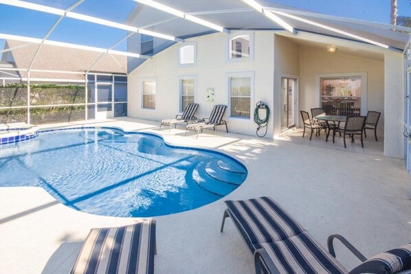 Una piscina con sillas de descanso, un porche cubierto y una zona de comedor con mesa y sillas.