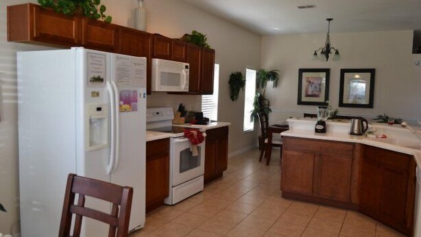 A kitchen with wooden cabinets, a white refrigerator, a microwave, and a stove. There is a dining table with chairs and a chandelier. - Ip60393 - The Shire at West Haven - 4 Bed 3 Baths Villa (Davenport)