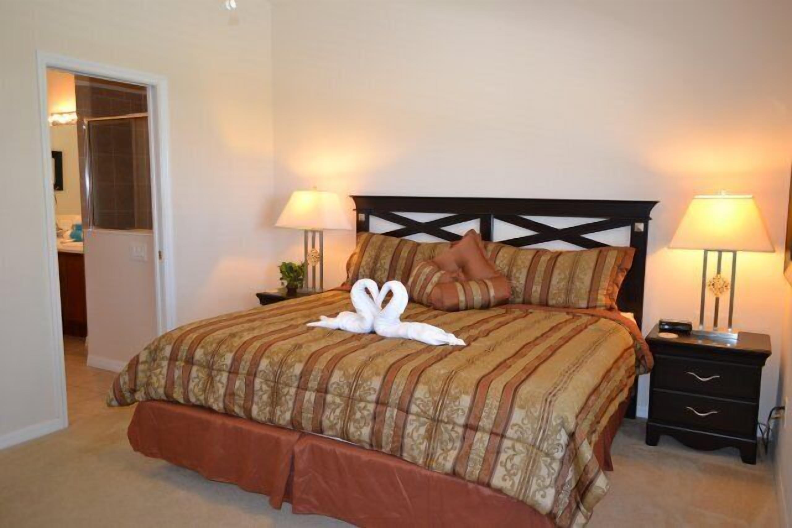 A neatly made bed with a brown and beige striped bedspread, two pillows, and a swan-shaped towel arrangement. There are two bedside tables with lamps, and a door leading to another room.