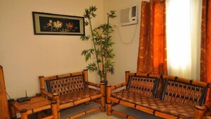 A living room with two bamboo sofas, a potted plant, a framed picture on the wall, and a window with orange curtains. - Haus Of Tubo Pension House (Davao)