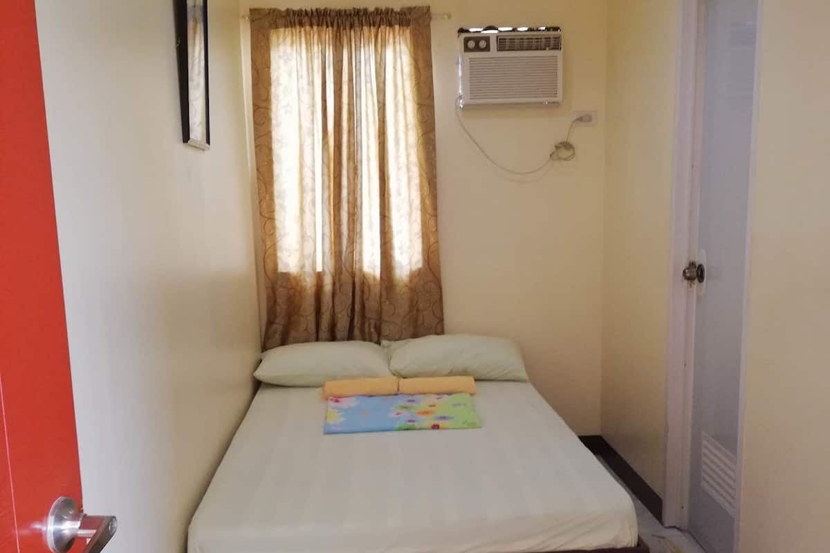A bedroom with a bed, a window with curtains, an air conditioning unit, and a door.