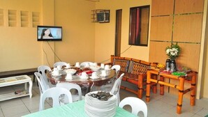 A dining area with a round table set for a meal, surrounded by white chairs. A television is mounted on the wall, and there is a wooden bench with cushions against the wall. - Haus Of Tubo Pension House (Davao)
