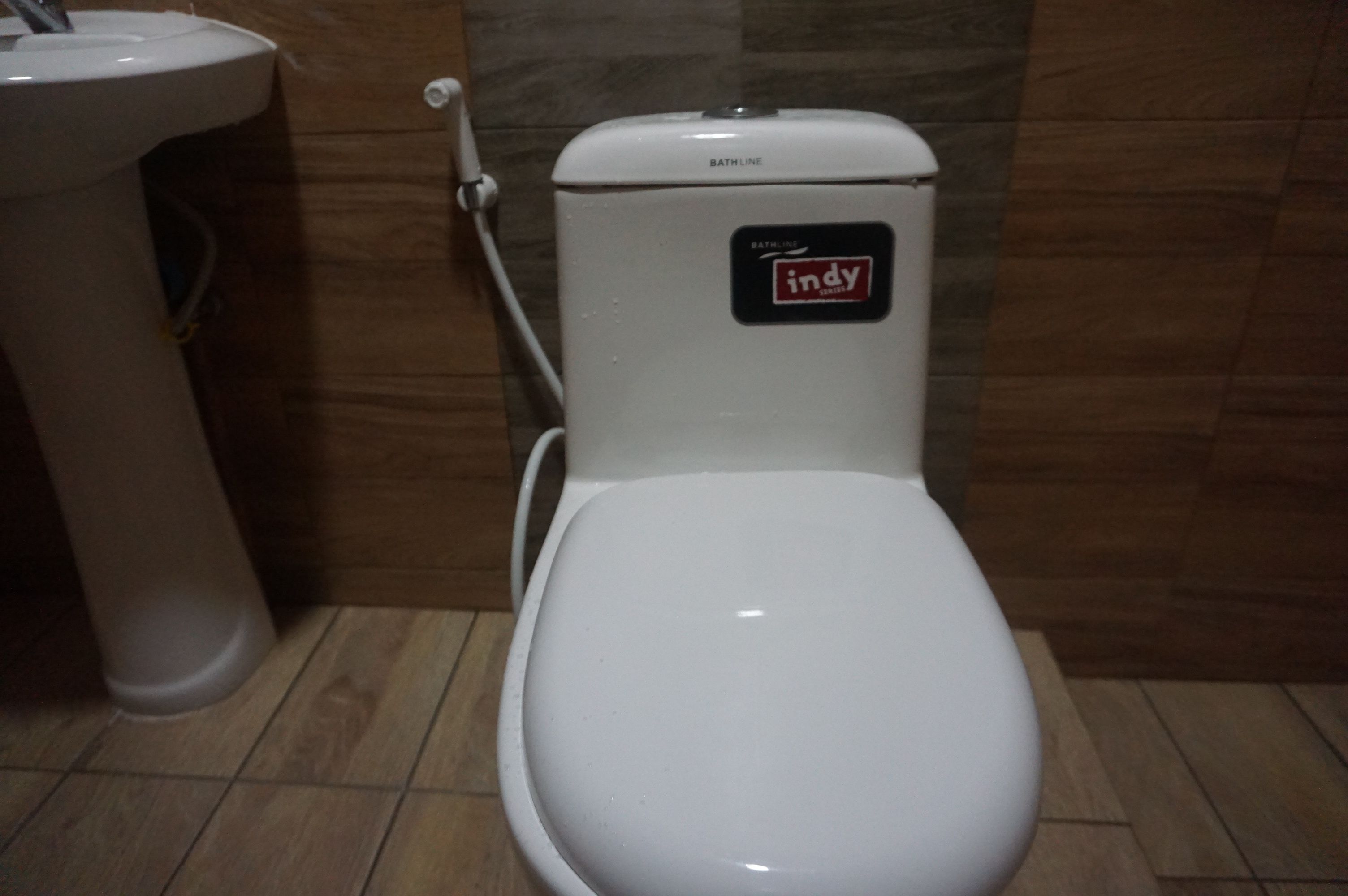 A white toilet with a 'Bathline' label and an 'Indy' sticker, positioned next to a pedestal sink in a bathroom with wooden flooring.
