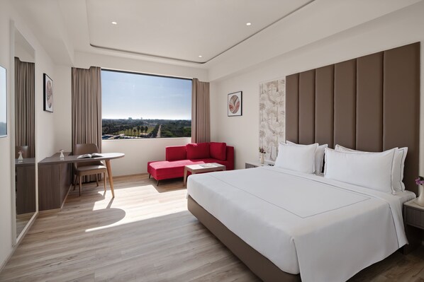 A modern hotel room with a large bed, a desk, a red sofa, and a view of the city. - Treat Resort Rajkot (Rajkot)