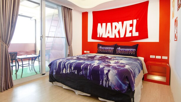 Un dormitorio con una cama, un gran cartel rojo con la palabra 'MARVEL' en la pared, y vistas al exterior a través de un ventanal corredero.