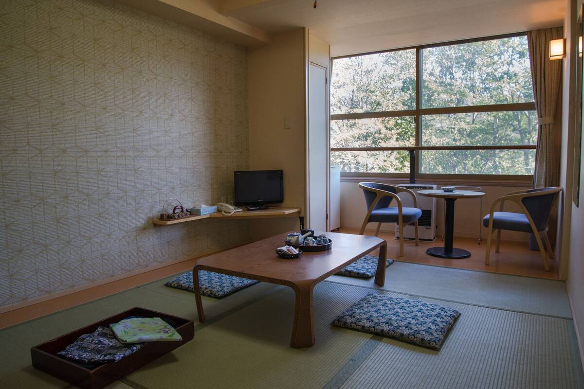 A traditional Japanese-style room with tatami flooring, a low table, chairs, a television, and a window with a view of trees.