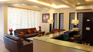 A modern living room with a glass coffee table, two brown sofas with orange cushions, a side table with a vase of flowers, and a desk with a computer and phone. - Parkwood Suites (Bengaluru)