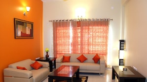 A living room with a sofa, coffee table, and a small table, with a patterned curtain and a framed picture on the wall. - Parkwood Suites (Bengaluru)