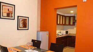 A dining area with a glass table, chairs, and orange placemats. There are framed pictures on the wall and a kitchen in the background. - Parkwood Suites (Bengaluru)