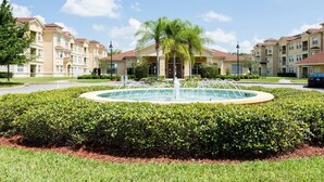 Un patio con una fuente circular, rodeado de setos cuidados y una palmera.