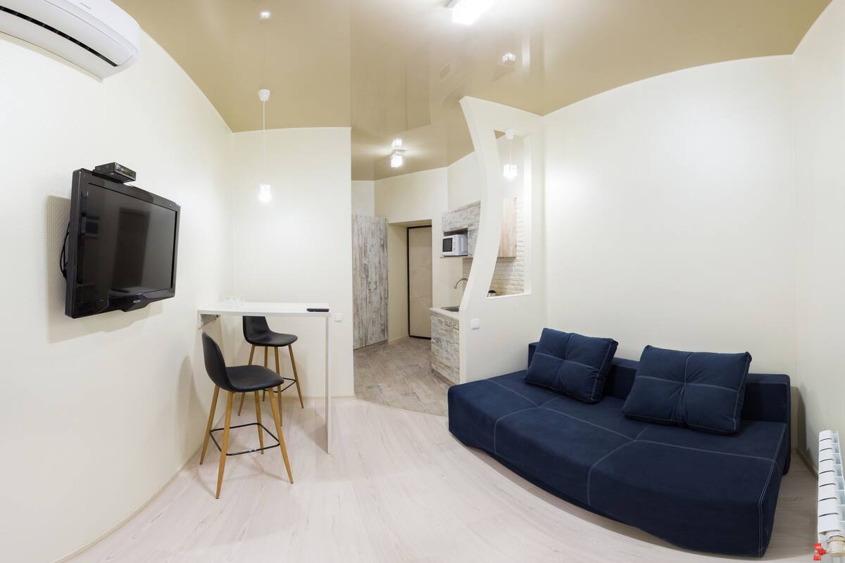 Una sala de estar moderna con un sofá azul oscuro, un televisor de pantalla plana montado en la pared y una pequeña zona de comedor con dos sillas.
