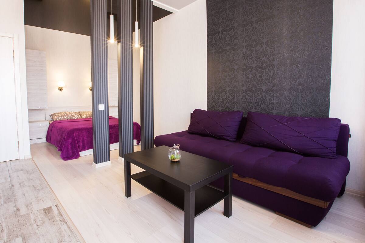 Una sala de estar moderna con un sofá morado, una mesa de centro oscura y una cama con cubrecama morado.