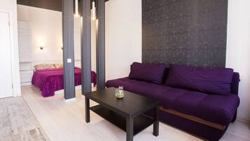 Un salon moderne avec un canapé violet, une table basse sombre et un lit recouvert d’une couverture violette.