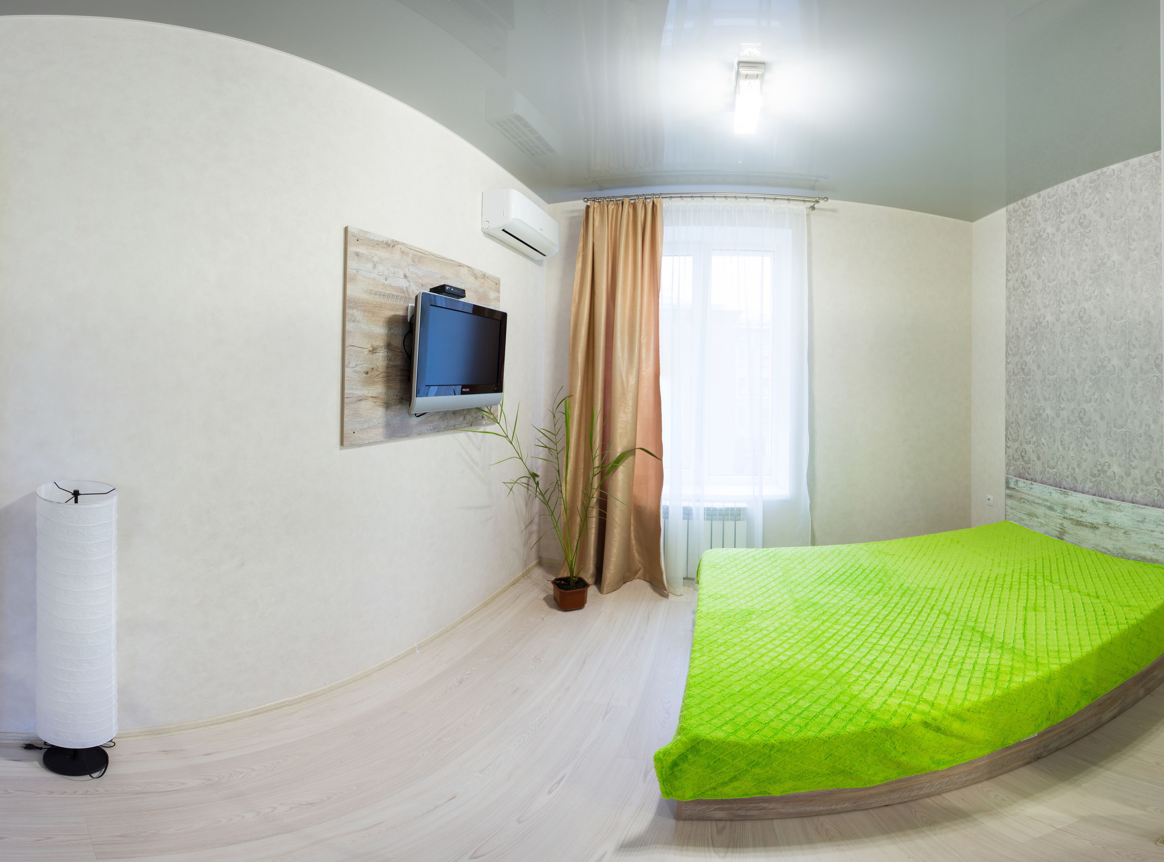 Un dormitorio con techo curvo, una cama con colchón verde, un televisor instalado en la pared, una planta en maceta y una ventana con cortinas.