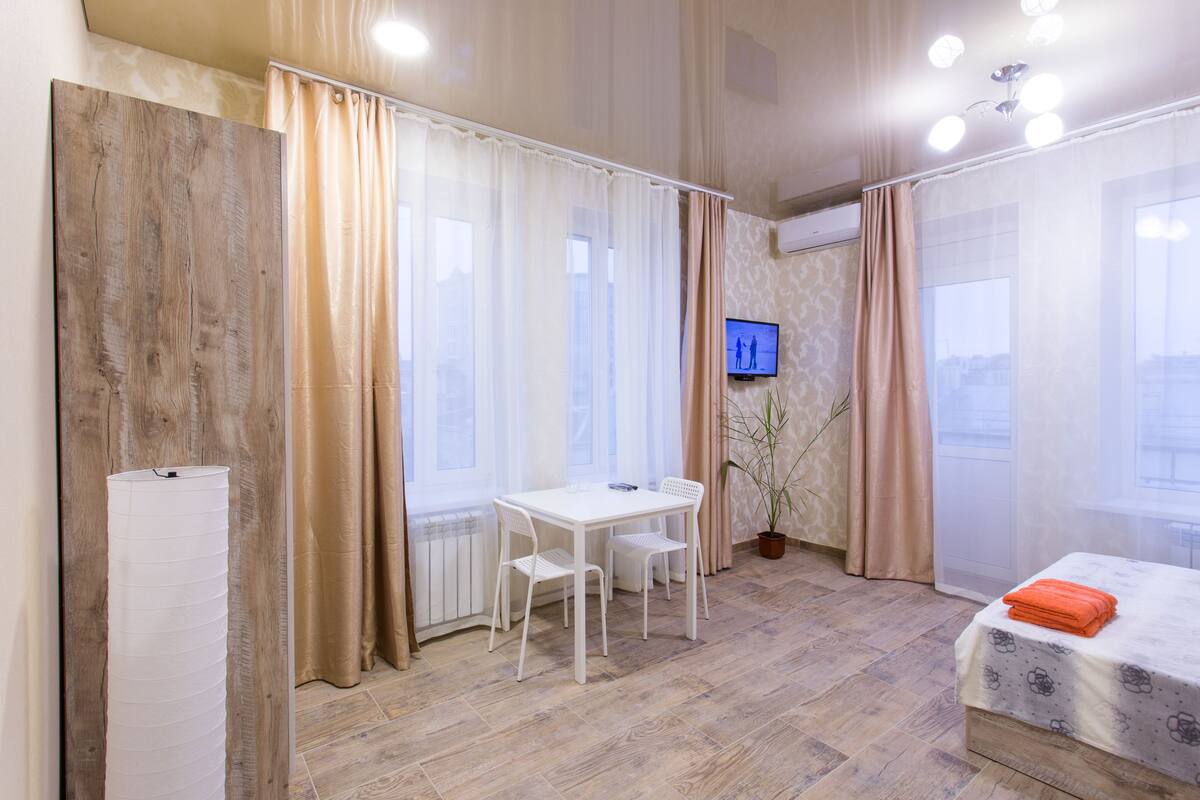Habitación de hotel con piso de madera, una mesa blanca con dos sillas, una cama y una toalla naranja, además de un televisor en la pared.