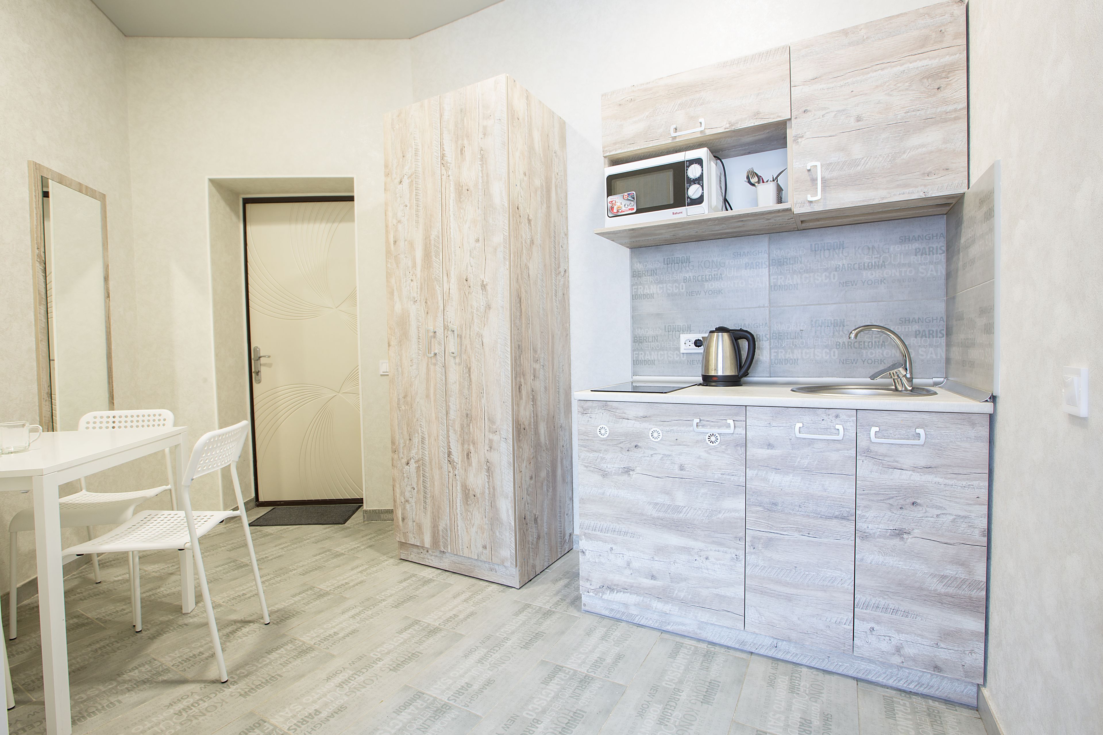 A compact kitchen with a white countertop, a microwave, and a sink. There is a white chair and a door leading to another room.