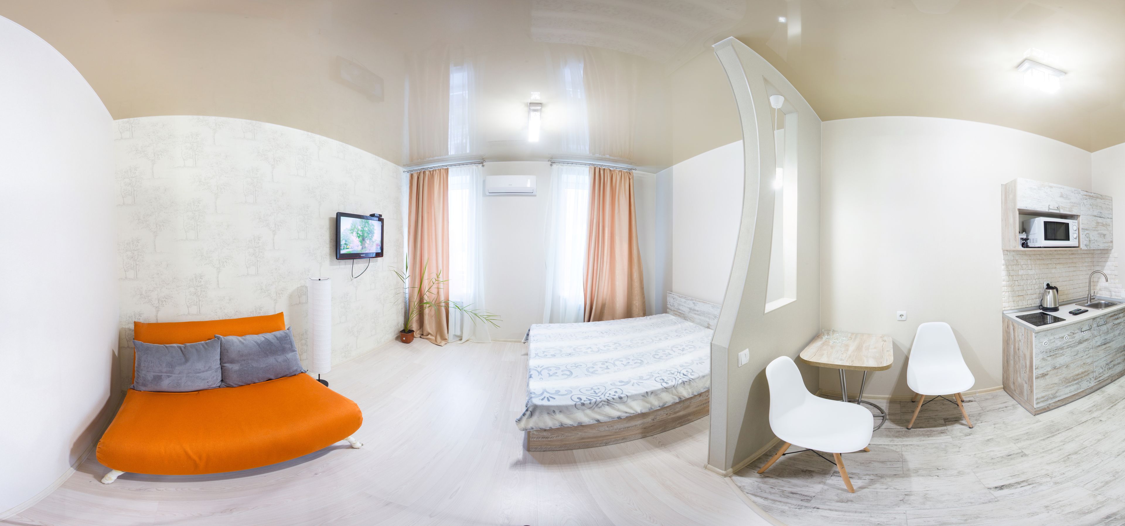 A modern hotel room with a curved bed, a small desk with a chair, a TV, and a kitchenette.